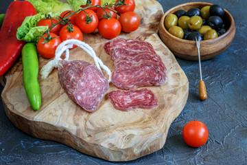 Sliced fresh fuet on the kitchen cutting board. Olives in wooden bowl and fresh vegetables. Healthy mediterranean food.