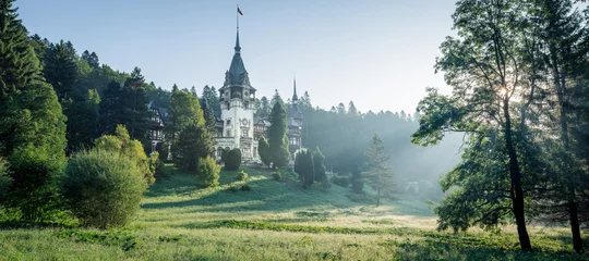 Fotobehang Blauwe hemel Peles Castle, famous residence of King Charles I in Sinaia, Romania. Peaceful summer landscape at sunrise of royal palace and park with mist and sun rays shining through the branches.  © Matteo Gabrieli