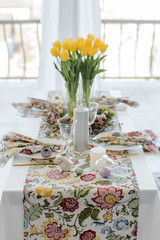 Festive decoration. Table with a floral tablecloth with linen napkins. golden cutlery, yellow tulips, wreath, happy easter concept.