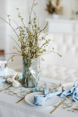 Decorative festive table setting with sky blue textile napkin and floral tablecloth, easter eggs, dishes and golden cutlery, glasses. willow in vase. holiday home interior.