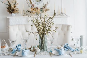 Decorative festive table setting with sky blue textile napkin and floral tablecloth, easter eggs, dishes and golden cutlery, glasses. willow in vase. holiday home interior.