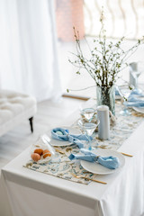 Decorative festive table setting with sky blue textile napkin and floral tablecloth, easter eggs, dishes and golden cutlery, glasses. willow in vase. holiday home interior.