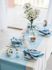 Decorative festive table setting with sky blue textile napkin and tablecloth, eggs, floral dish and golden cutlery, glass for wine. Happy easter concept.