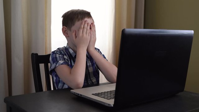 4k. a Surprised 10-year-old child looks at the computer screen with a shocked and scared look, covering his eyes with his hands while watching a scary movie. Danger of the Internet.