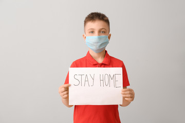 Little boy with protective mask holding paper with text STAY HOME on grey background. Concept of...