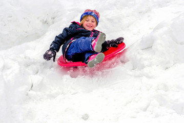Fototapeta na wymiar Laughing child sleds down a snowy hill on a red saucer sled 