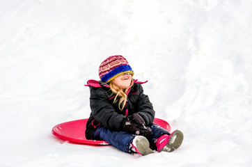 Fototapeta na wymiar Laughing girl with hat over her eyes, sits on a red saucer sled as she plays on a snowy winter day