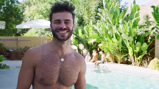 Portrait of smiling bare chested Hispanic man standing by swimming pool with group of friends enjoying summer pool party together - shot in slow motion