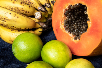 Close up to a fresh sliced orange papaya with seeds inside, a freckled bananas cluster and three green lemons over a blue navy background. Fruit and eating concept photography