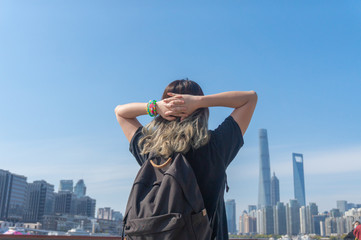 Fototapeta na wymiar Young asian girl with dyed gray curvy hair and bracelet holding hands behind her head and have a rest while travelling in the background of Shanghai cityscape and the blue sunny sky