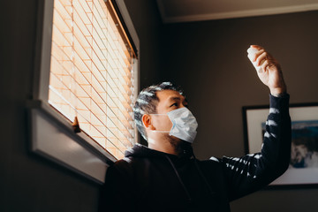 Asian man in isolation looking at medicine bottle. Man in medical mask forced to stay inside the...