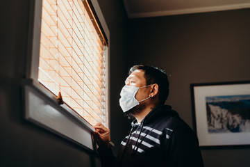 Asian man in isolation looking through window blinds. Man in medical mask forced to stay inside the...
