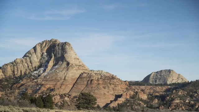 A golden hour timelapse of red cliffs in Zion National Park lit with pleasant sunlight. Clouds flow overhead.