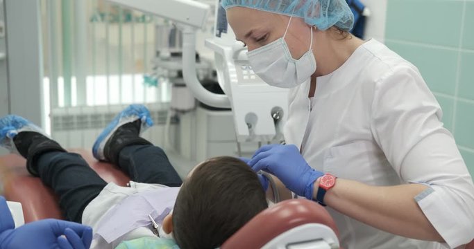 Nice dentist doctor is doing dental treatment for his little patient. Doctor complies with all safety standards, she wears uniform, medical mask, gloves and uses sterile medical instruments. Portrait