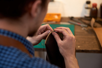 Closeup of craftsman hands sewing brown leather parts with needle for future case for glasses. The man with needle sewing leather in the workshop. 