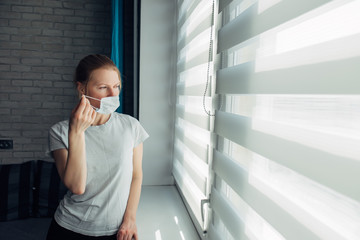 Young woman in mask looking through the blinds. Quarantined coronavirus person. Stay home. Stop the pandemic. Self-isolation, putting up mask.