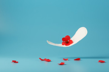 Sanitary pad in flight on a blue background with fallen petals of red flowers. Concept of the beginning of menopause.