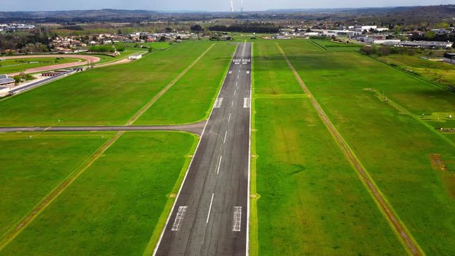 Aerial view over a landing runway with a drone which is going lower and lower in the on the runway. There is green grass around the empty airstrip without airplane, with a cloudy sky - 4K