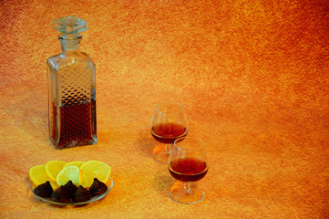 Two glasses and a crystal bottle with cognac stand next to a plate with lemon and chocolate candies...