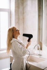 Beautiful women enjoying in hotel room. Young happy smiling woman wearing bathrobe using hair dryer in bathroom.	