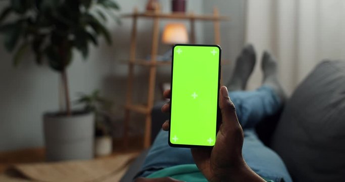 Young man lying on couch with modern smartphone in his hand while looking on mock up screen with trecking markers on it. Concept of green screen and chroma key. Cosy home background.