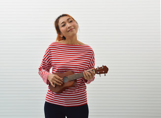 Happy young asian woman wearing red and white stripped shirt playing ukulele guitar standing over white wall background, Smiling facial expression - Powered by Adobe