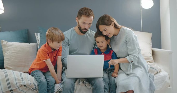 Young parents with small children watching video on laptop sitting on couch. Mother, father and two little sons surfing internet on laptop relaxing on couch at home on quarantine