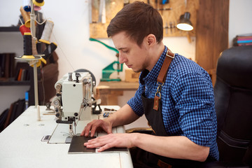 The man behind white sewing machine. The photo of the room with white table, sewing machine on it and working man. Manufactory and small business of leatherman. 