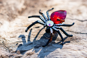An extravagant women's brooch in the form of a black spider with rhinestones and a red stone, shimmering in the sunlight under the open sky on a wooden surface. Red forest ants attack the spider - Powered by Adobe
