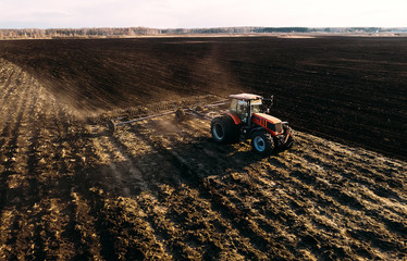 Aerial view of tractor plows the field in sunset, sunrise, raising dust, and behind it fly birds. Farm tractor with harrow plow preparing land for sowing. Agriculture industry, cultivation of land.