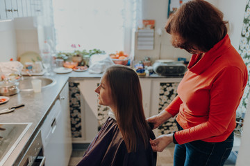 mature woman cuts hair to young beautiful woman with long hair at home, because of quarantine