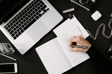 Designer hand writes in his daily planner. Workplace with laptop, vintage film camera, pafume,...