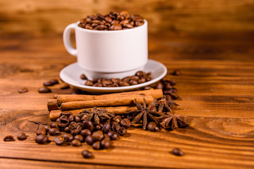 White cup filled with coffee beans, star anise and cinnamon sticks on wooden table