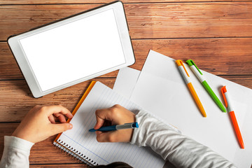 Top view tablet with blank screen on wooden table in front of boy writing in a notebook next to...