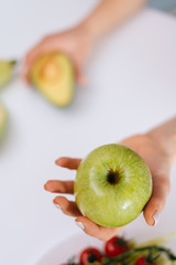 Close-up of green tasty apple in woman hand on white background, avocado on table. Concept of healthy eating.
