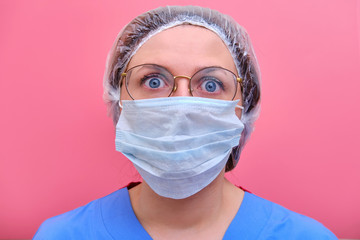 Portrait of a doctor woman with a frightened facial expression, closeup. Nurse in a blue uniform and a medical mask on a pink background.