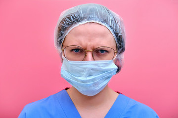 Portrait of a doctor woman with angry facial expression, close up. Nurse in a blue uniform and a medical mask on a pink background. Stay at home.