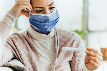 Worried woman with face mask measuring temperature at home.