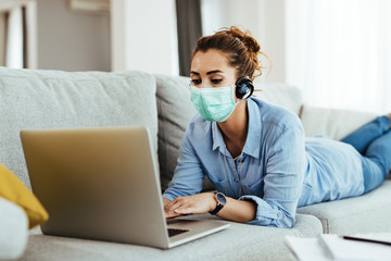 Young woman with face mask using laptop while relaxing on the sofa.