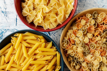 blue, red and yellow ceramic bowls with different pastas on a white and blue textured background