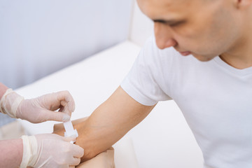 Nurse in rubber protective gloves putting an adhesive bandage on young man's arm vein after blood test or injection of vaccine, close-up. Concept of healthcare and medicine.