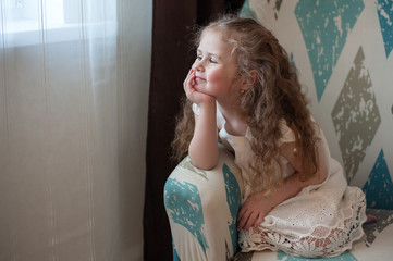 Fototapeta premium Sad little girl sitting in a chair and looking longingly out the window