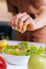 Close-up of women's hands squeezing juice from fresh yellow lemon into salad bowl with sliced vegetables. Red bell pepper, avocado, lemon on the table. Concept of healthy food lifestyle.