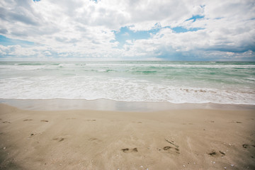Human footprints go into the ocean waters on the beach. The water is azure, and there are many beautiful clouds.