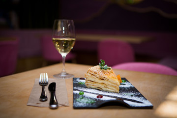 On the table is a glass of white wine and a portion of delicious cake on a black plate sprinkled with powdered sugar, next to which are appliances for food.