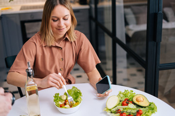 Young pretty woman mixing fresh vegetable salad while sitting at table in kitchen with modern interior, mobile phone on hand. Concept of healthy eating.