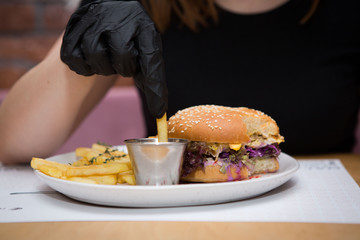 A girl in a black T-shirt and black latex gloves dips french fries in ketchup, she has a burger with meat on a plate.