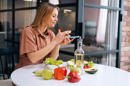 Beautiful young woman uses smartphone for taking photos of vegetables lying on kitchen table. Concept of social networks. Concept of healthy eating.