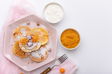 pancakes with raspberries and physalis, powdered sugar and maple syrup in pink plate with sour cream and mango jam on white background