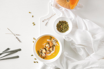 Pumpkin soup, a piece of pumpkin, seeds lie on a white table. Nearby is a white towel and serving utensils. 
